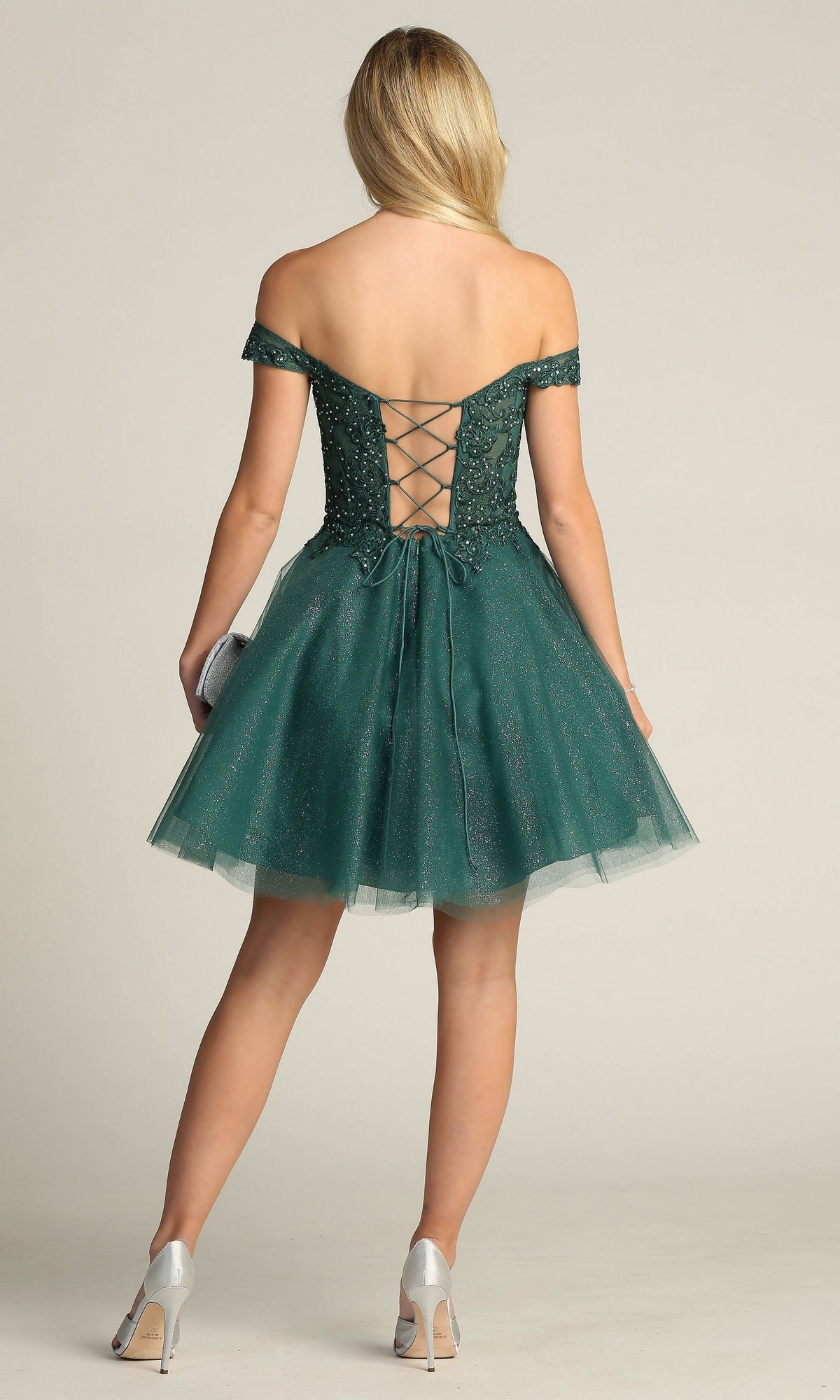A woman wearing the Off-Shoulder Short Glitter Homecoming Dress C318 with a lace-up back stands facing away, holding a clutch and wearing silver heels.